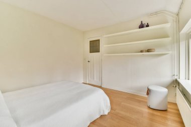 Parlak, minimalist bir yatak odası, beyaz örtülü bir yatak odası, ahşap döşemeler, ve sakin atmosferi arttıran basit raflar..