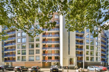 Çok sayıda balkon ve geniş pencereli, yemyeşil ağaçlar ve açık bir gökyüzünün arasına kurulmuş çağdaş bir konut binası..