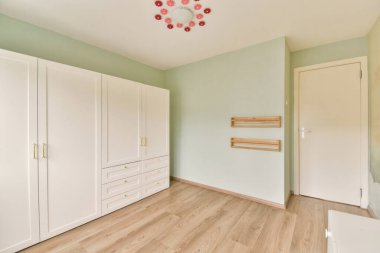 Açık yeşil duvarlar, ahşap döşemeler ve minimalist depolama çözümlerinin yer aldığı geniş bir oda, huzurlu ve çağdaş bir yaşam alanı yaratıyor..