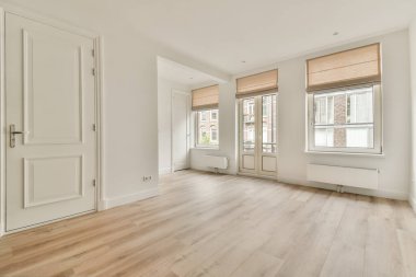 Geniş, modern bir oturma odası. Açık renkli duvarları, ahşap döşemeleri ve minimalist perdelerle kaplı büyük pencereleri var..