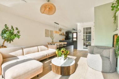 Rahat bir kanepe, dekoratif bir sehpa ve canlı bitkilerin olduğu parlak ve modern bir oturma odası. Güneş ışığı içeri akar ve sıcak bir atmosfer yaratır..