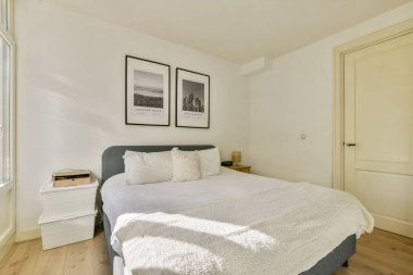 Zarif, modern bir yatak odası yumuşak çarşaflarla süslenmiş bir yatak odası, duvarda çerçevelenmiş sanat baskıları ve doğal ışığın yarattığı rahat bir ortam..