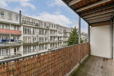 Modern bir apartmana bakan geniş bir balkon, içinde birden fazla balkon ve yeşil ağaçlar var. Gökyüzü açık, sakin bir atmosfer yaratıyor..