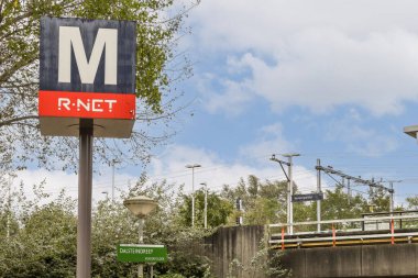 R-NET hattını gösteren bir tren istasyonu tabelası mavi gökyüzü ve arka planda yeşillik, kentsel ulaşım özelliklerini gösteriyor..