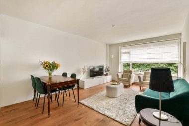 İçinde ahşap bir yemek masası, yeşil sandalyeler ve büyük pencerelerden gelen doğal ışık bulunan modern bir yaşam alanı. Odanın zarif ve rahat bir atmosferi var..