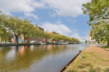 Huzurlu bir kanal sahnesi, bankalar boyunca canlı yeşillikler, modern mimari ve açık bir gökyüzü eşliğinde, huzurlu bir şehir manzarası yaratıyor..