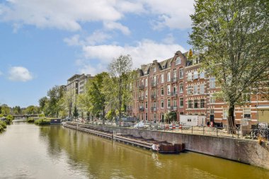 Ağaçlarla ve tarihi binalarla kaplı bir kanal manzarası. Canlı bir şehir manzarası, tekneleri ve açık mavi gökyüzü..