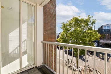 Güneşli bir balkon, dışarıda modern mimari tarafından çerçevelenmiş canlı bir ağaç ve aşağıdaki hareketli bir sokak manzarasını gözler önüne seriyor..