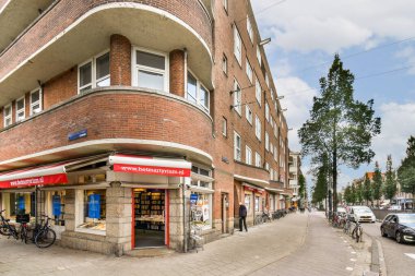 Perakende alanları, yayaları ve kaldırım boyunca parkedilmiş bisikletleri olan modern bir binayı, parçalı bulutlu bir gökyüzünün altında gösteren canlı bir sokak manzarası..