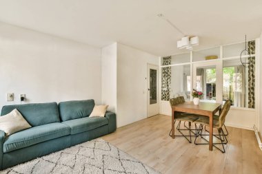 Günümüz oturma odasında açık renkli bir kanepe, ahşap yemek masası ve geniş pencerelerden yayılan doğal ışık, hoş bir atmosfer yaratıyor..