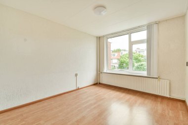 Geniş pencereli, parlak ve ferah boş bir oda. Doğal ışığın içeri akmasına izin veren, basit ahşap zemin ve nötr duvarları aydınlatan..