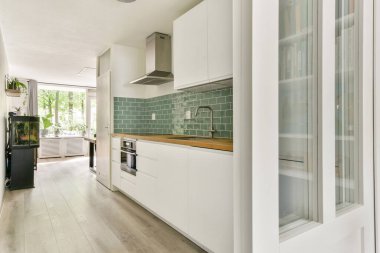 Beyaz dolaplar, ahşap aksanlar ve yeşil fayanslardan oluşan gösterişli, modern bir mutfak. Doğal ışık geniş pencerelerden süzülerek parlak bir atmosfer yaratır..