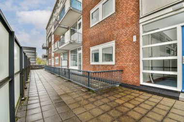 Çağdaş bir apartman binası. Tuğladan yapılmış bir cephesi ve ona uzanan kaldırımlı bir yolu var. Arka planda açık bir gökyüzü var..
