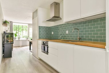 Beyaz dolaplar, ahşap bir tezgah ve yeşil fayanslı duvarlar içeren gösterişli bir mutfak, büyük pencereler ve kapalı bitkilerden gelen doğal ışıkla tamamlanmış..