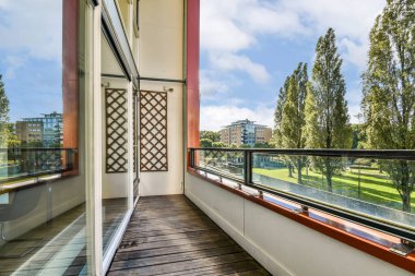 Gösterişli, iyi tasarlanmış bir balkon alanı, arka planda yapraklı ağaçlar ve modern binalarla dolu çarpıcı bir park manzarası sunuyor. Rahatlamak için ideal..