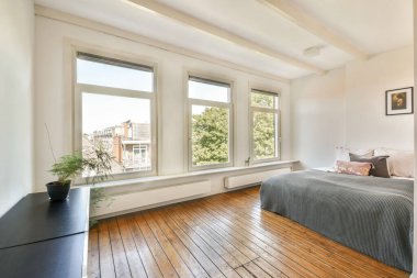 Doğal ışığı davet eden, rahat mobilyaları ve sıcak ahşap zemini vurgulayan geniş pencereli çağdaş bir yatak odası..