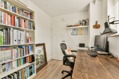 Ahşap bir masa, ergonomik sandalye ve çeşitli eşya ve kitaplarla dolu düzgün düzenlenmiş kitaplıklardan oluşan bir duvar içeren şık ve düzenli bir ev ofisi..
