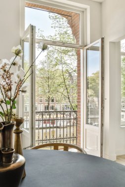 Zarif bir masa üzerinde dekoratif bir çiçek düzeniyle doğal ışığı davet eden açık pencereleri gösteren zarif bir iç mekan. Hoş bir atmosfer yaratıyor..