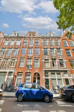 Resimli bir sokak manzarası, büyük pencereli ve park edilmiş arabalı zarif kırmızı tuğlalı binalar, Amsterdam 'da kentsel yaşamı simgeliyor..