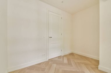 Minimalist bir iç mekanın parlak ve havadar bir köşesi açık renkli bir duvara karşı bir kapı sergiliyor, ringa kemiği desenli bir zemin ile tamamlanıyor..