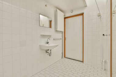 Pürüzsüz beyaz fayanslar, modern bir lavabo, temiz bir kapı tasarımı, minimalist estetiği ve işlevselliği simgeleyen geniş ve havadar bir banyo..
