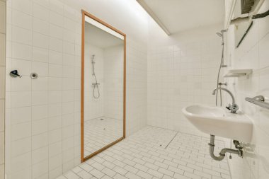 Geniş, minimalist bir banyo. Beyaz fayanslı duvarlar, zemin, büyük bir ayna ve parlak tesisat malzemeleri. Düzenli tasarım temiz ve huzurlu bir ortam sunuyor..