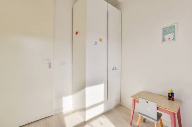 Beyaz bir gardırobu ve küçük bir masası olan minimalist bir çocuk odası. Çocuklar için rahat ve düzenli bir yer yaratmak için ideal..