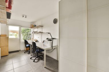 Doğal ışığı maksimuma çıkarmak için tasarlanmış, masaları, rafları ve yeşile bakan geniş bir penceresi olan modern bir ofis alanı..
