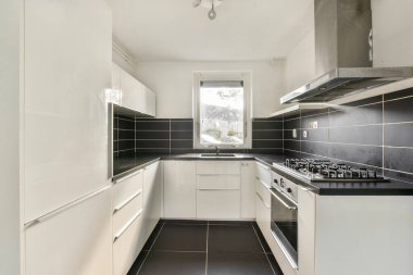 Bu mutfak, beyaz dolaplı, siyah tezgahlı ve modern aletli, işlevselliği ve tarzı sıkışık bir alanda vurgulayan parlak bir tasarıma sahiptir..