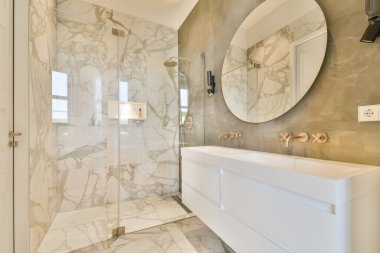 Zarif mermer duvarları, şık yuvarlak bir aynası ve zarif bir dolabı olan modern bir banyo sofistike bir atmosfer yaratıyor..