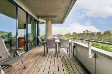 Gösterişli sandalyeler ve ahşap zeminden oluşan modern bir balkon, arka planda yeşil ağaçlar ve kentsel manzaralar sunuyor..