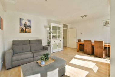 Duvarlarda rahat bir kanepe, yemek masası ve sanat eserlerinin bulunduğu, pencerelerden yansıyan güneş ışığıyla tamamlanmış, parlak ve havadar modern bir oturma odası..