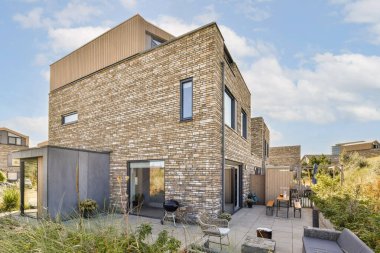 Teras mobilyası ve peyzajı olan yemyeşil bir bahçeye kurulmuş, tuğla kaplamalı ve geniş pencereli çağdaş bir konut yapısı..