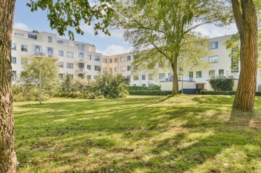 Arka planda uzun ağaçlar, çimenler ve modern yerleşim yerlerinin yer aldığı yemyeşil bir park, sakin bir kentsel çevre sağlıyor..