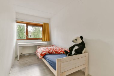 Parlak ve basit bir çocuk odası. İçinde ahşap bir yatak, renkli bir yatak ve pencerenin yanında sevimli bir panda oyuncağı var. Doğal ışık davetkar atmosferi güçlendirir.