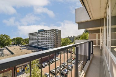 Geniş bir balkon manzarası modern binaları ve açık mavi gökyüzünün altındaki ağaçlar tarafından çerçevelenmiş hareketli bir otoparkı sergiliyor..