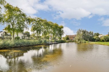 Sular boyunca canlı yeşilliklerin yer aldığı sakin bir göl manzarası, yukarıdaki berrak gökyüzünü dinlenmek için mükemmel bir ortamda yansıtıyor..