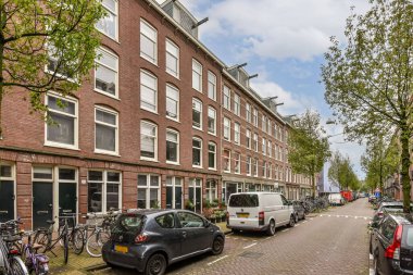 Çizgili binaların, park edilmiş bisikletlerin ve parlak bir gökyüzünün altındaki ağaçların yer aldığı pitoresk bir sokak sahnesi, kentsel yaşamı ve sükuneti gözler önüne seriyor..