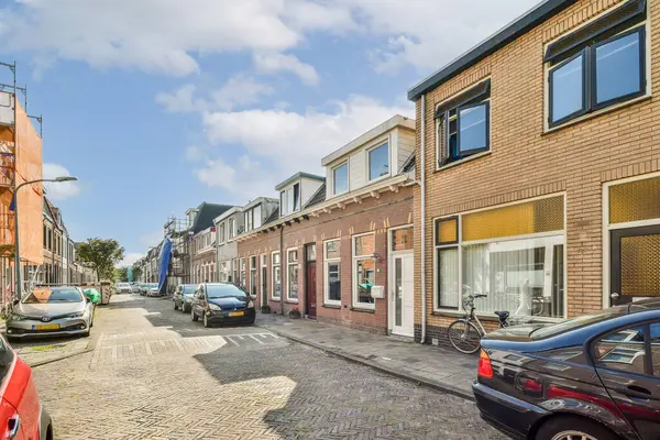 Parlak güneş ışığı ve dağınık bulutlarla aydınlatılan konut binaları ve park edilmiş arabalarla dolu sakin bir şehir caddesinin manzarası..