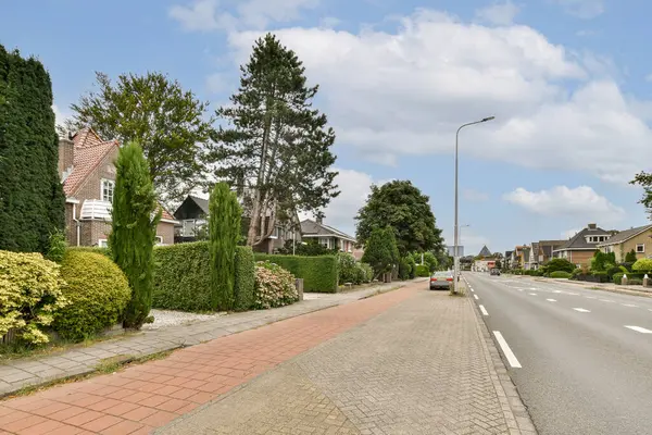 Parça bulutlu bir gökyüzü altında huzurlu bir banliyö ortamını gösteren, iyi korunmuş yeşil bitki ve ağaçlarla dolu sakin bir yerleşim yeri..