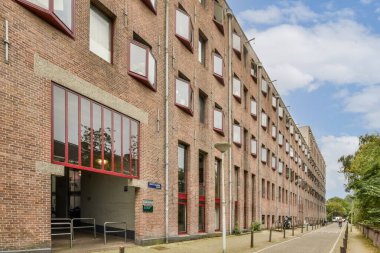 Sokak lambaları ve yeşilliklerle kaplı asfalt bir yol ile çevrili çok sayıda pencereye sahip çağdaş bir tuğla yapı..