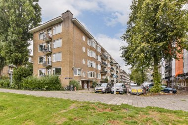 Ağaçlar ve otlarla çevrili modern bir yerleşim yeri manzarası, doğa ve şehir yaşamının bir karışımını sergiliyor. Mimaride büyük pencere ve balkonlar yer alıyor..