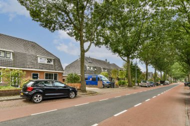 Yapraklı ağaçlarla kaplı sakin bir banliyö caddesi, konut evleri ve park edilmiş arabalar sergileniyor, sakin bir mahalle atmosferini somutlaştırıyor.