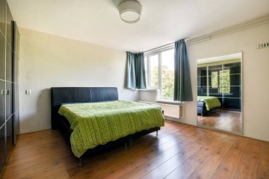 Geniş, modern bir yatak odası yeşil desenli yatak örtüsü, bir ayna ve doğal ışığa izin veren büyük pencereli..