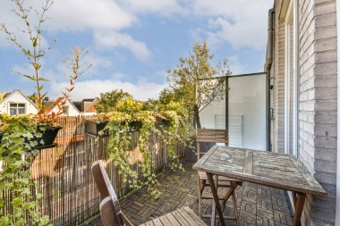 Bereketli bitkilerle süslenmiş sakin bir balkon ve berrak mavi gökyüzüne karşı kurulmuş ahşap bir masa kentsel bir ortamda huzurlu bir geri çekilme sunuyor..