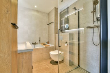 Geniş ve zarif tasarlanmış modern bir banyo. İçinde cam bir duş, parlak bir küvet ve modern eşyalar var. Huzurlu bir ortam yaratıyor..