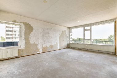 Bitmemiş duvarlar ve açık hava manzarası sunan geniş pencerelerle dolu boş bir oda. Zemin çıplak çimento, minimalist alanı vurguluyor..