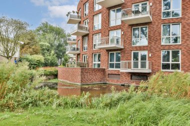 Çağdaş tuğladan bir apartmanda geniş pencere ve balkonlar, yemyeşil alanlar ve sakin bir atmosfer için huzurlu bir su yolu bulunur..