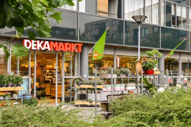 Dışarıdaki çiçeklerin ve bitkilerin olduğu canlı bir market cephesi. Parlak tabela, girişi çevreleyen modern mimari ve yeşillikle çelişiyor..