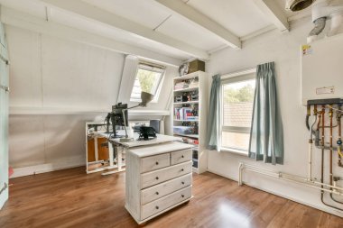 Geniş ve iyi ışıklandırılmış bir dikiş odası. Çalışma istasyonu, malzeme dolu raflar ve geniş pencerelerden yayılan doğal ışık..